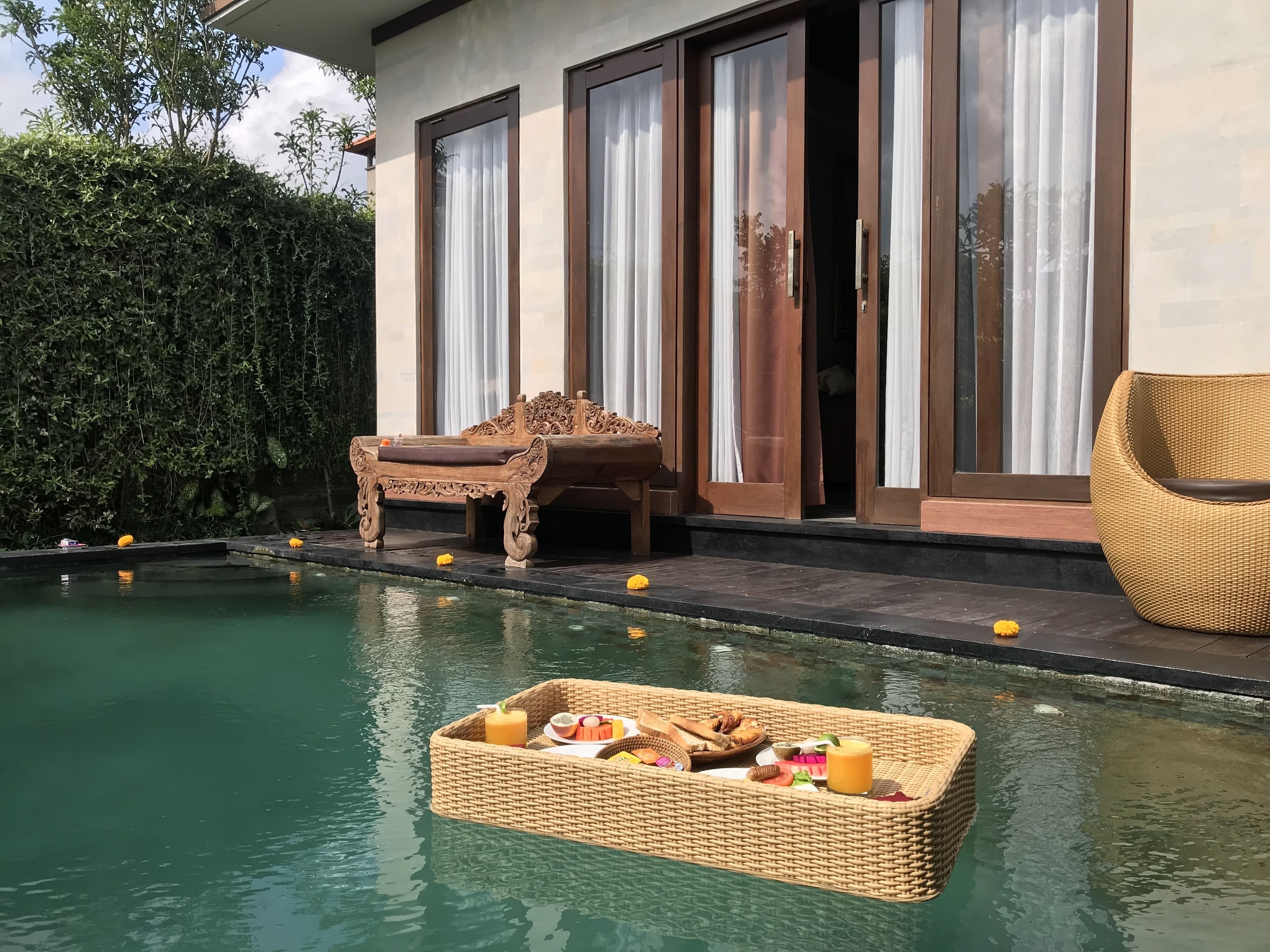 A pool area with a floating basket of food, a bench, and a chair near a building with large windows and a hedge.