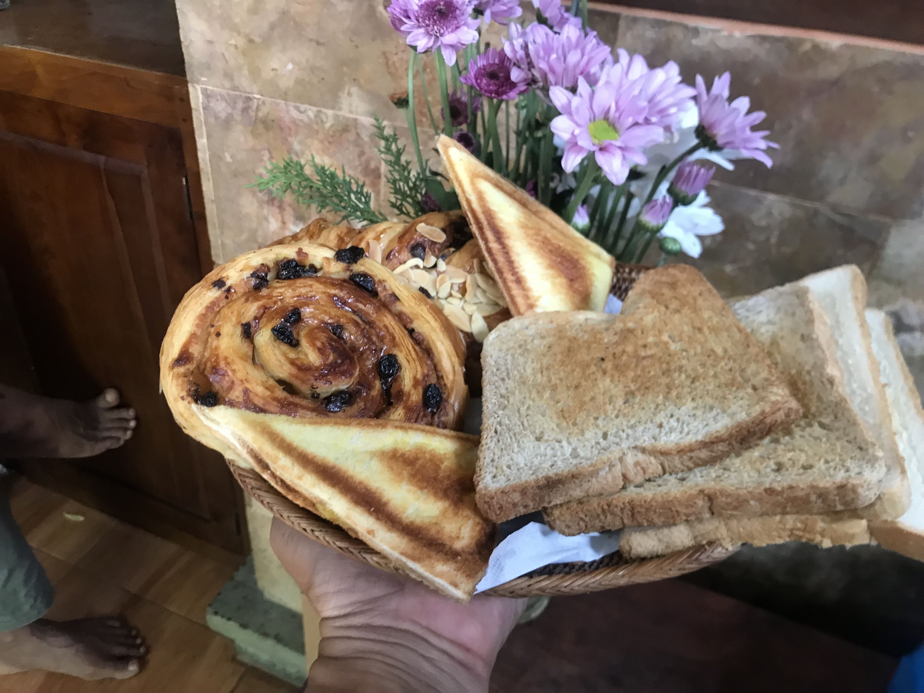A hand holding a basket with a cinnamon roll, sliced bread, and a bouquet of flowers.