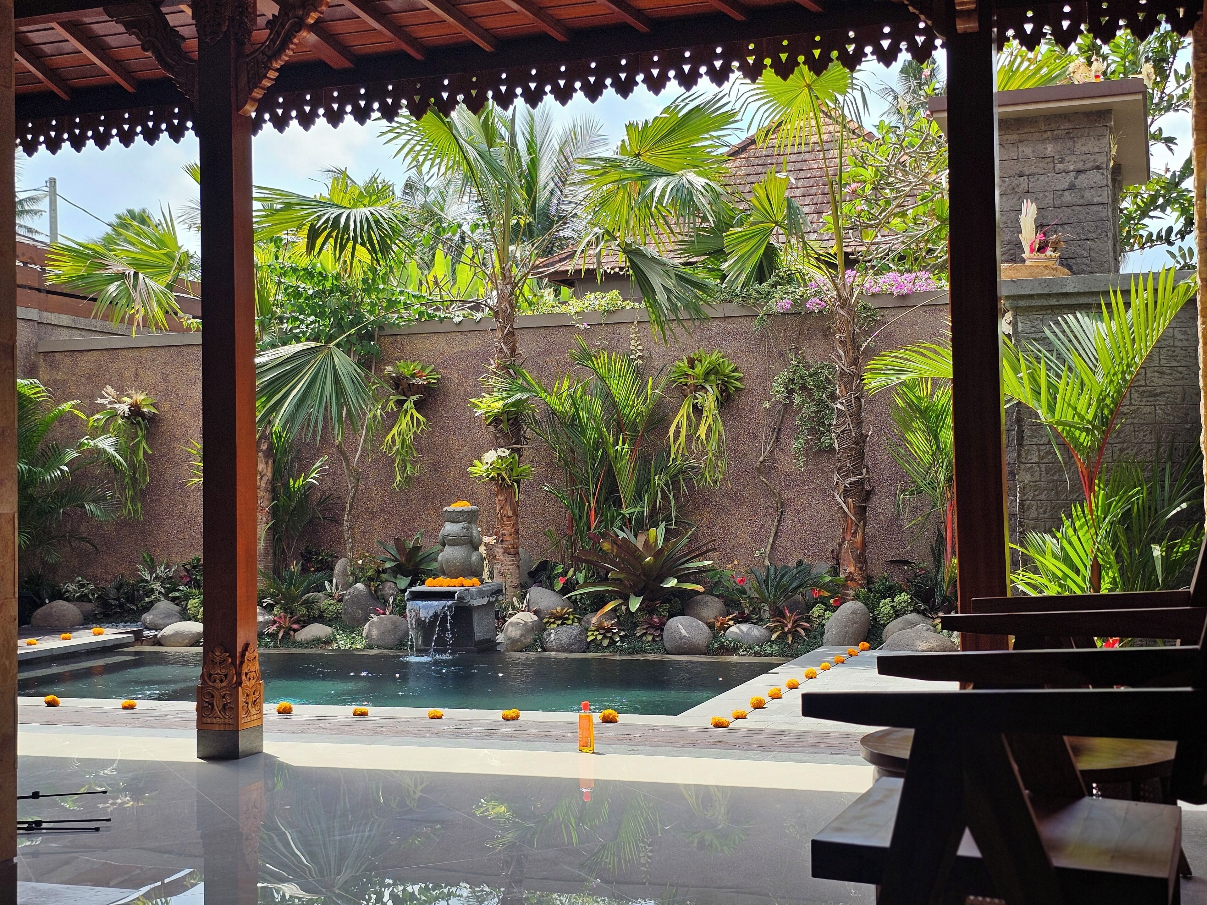 A covered outdoor area with a pool, surrounded by tropical plants and a stone wall.