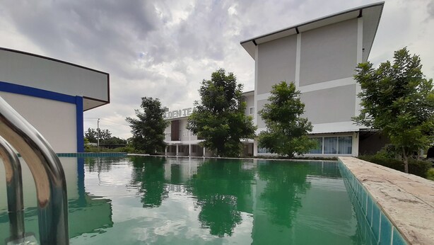 A swimming pool with a clear blue water surface, surrounded by a stone deck and metal railings. In the background, there are modern buildings with large windows and a covered area. - Golden Teak Ville (Buriram)