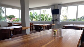 A spacious dining area with wooden tables and chairs, large windows with curtains, and a television mounted on the wall. - Golden Teak Ville (Buriram)