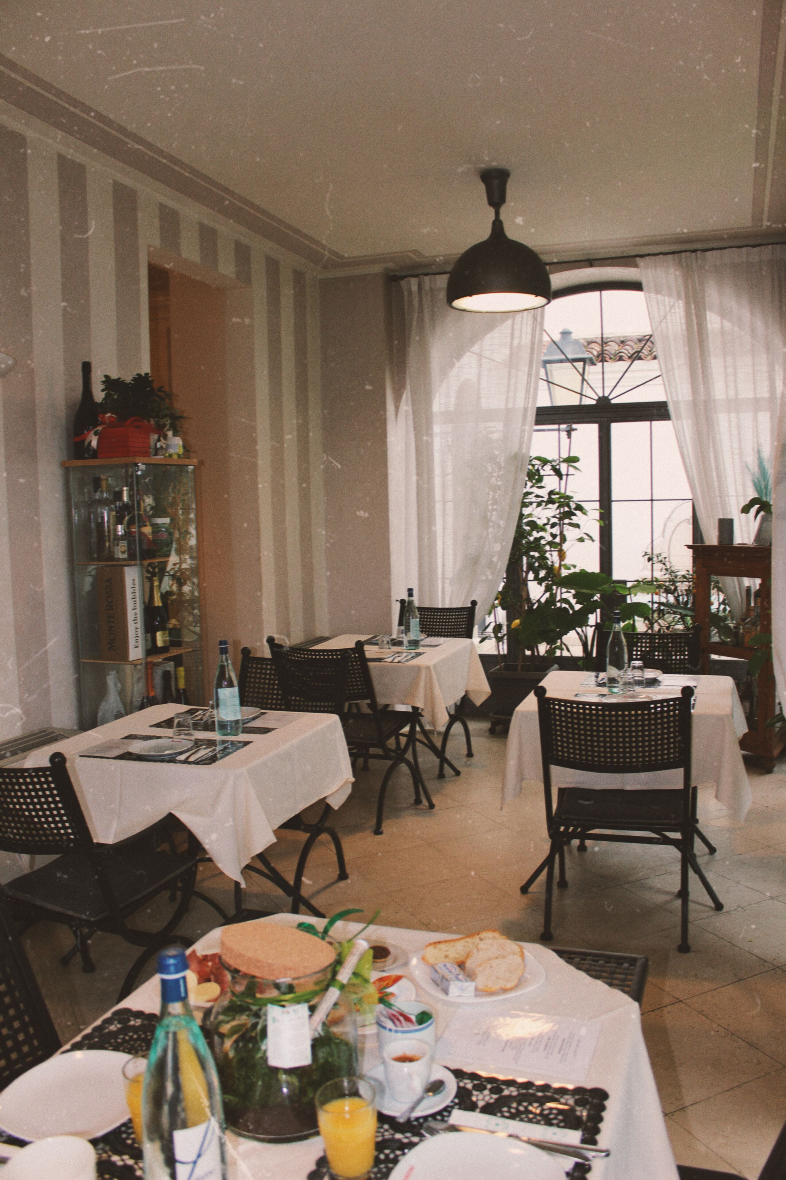 A dining area with tables set for meals, chairs, a chandelier, and a view through a window with curtains.