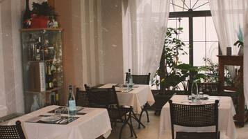 A dining area with tables set for meals, chairs, a chandelier, and a view through a window with curtains.