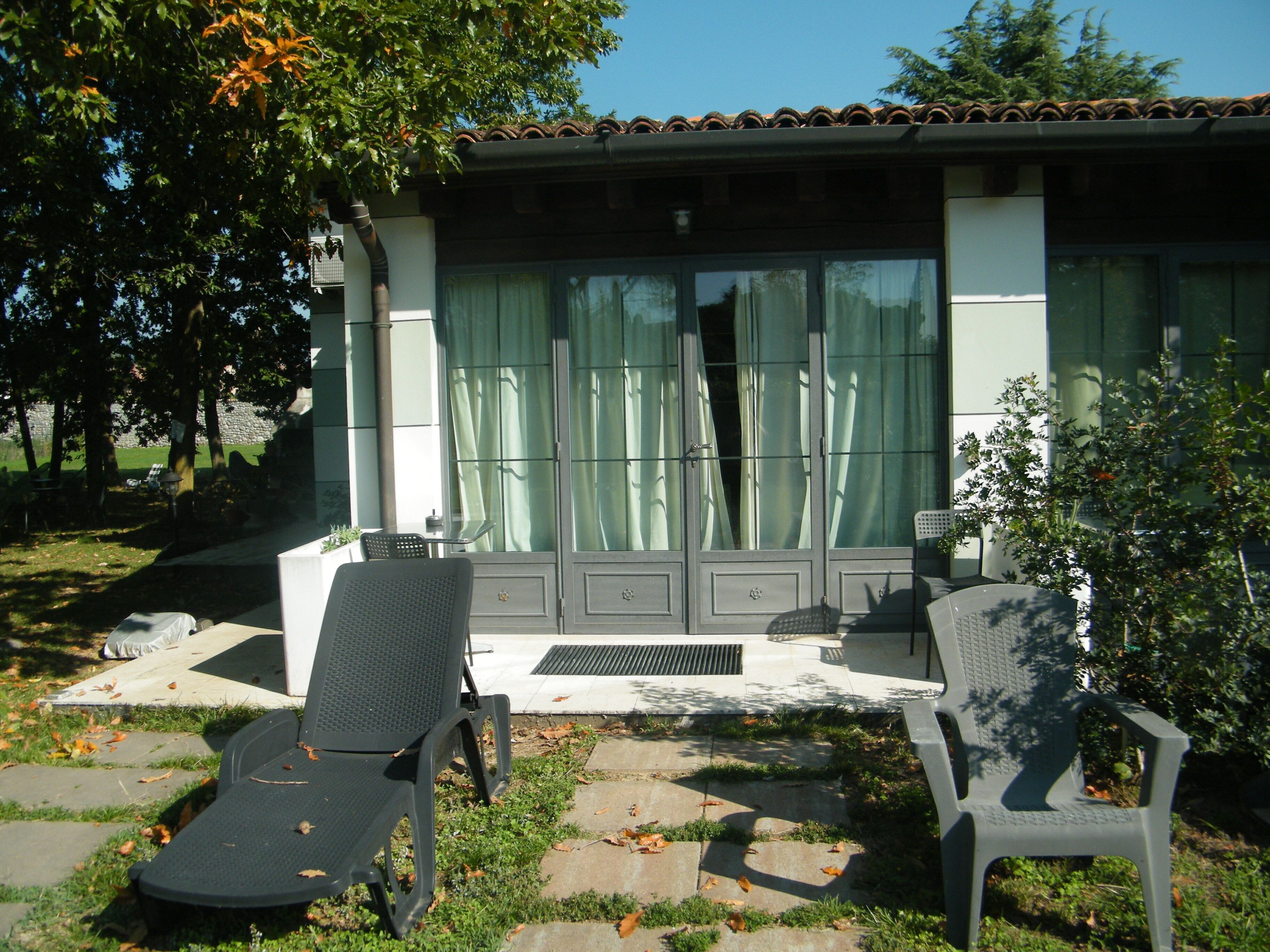 Ein Terrassenbereich mit zwei Liegestühlen und einem kleinen Tisch, ein Ziegeldach und ein Gebäude mit großen Fenstern und Vorhängen.