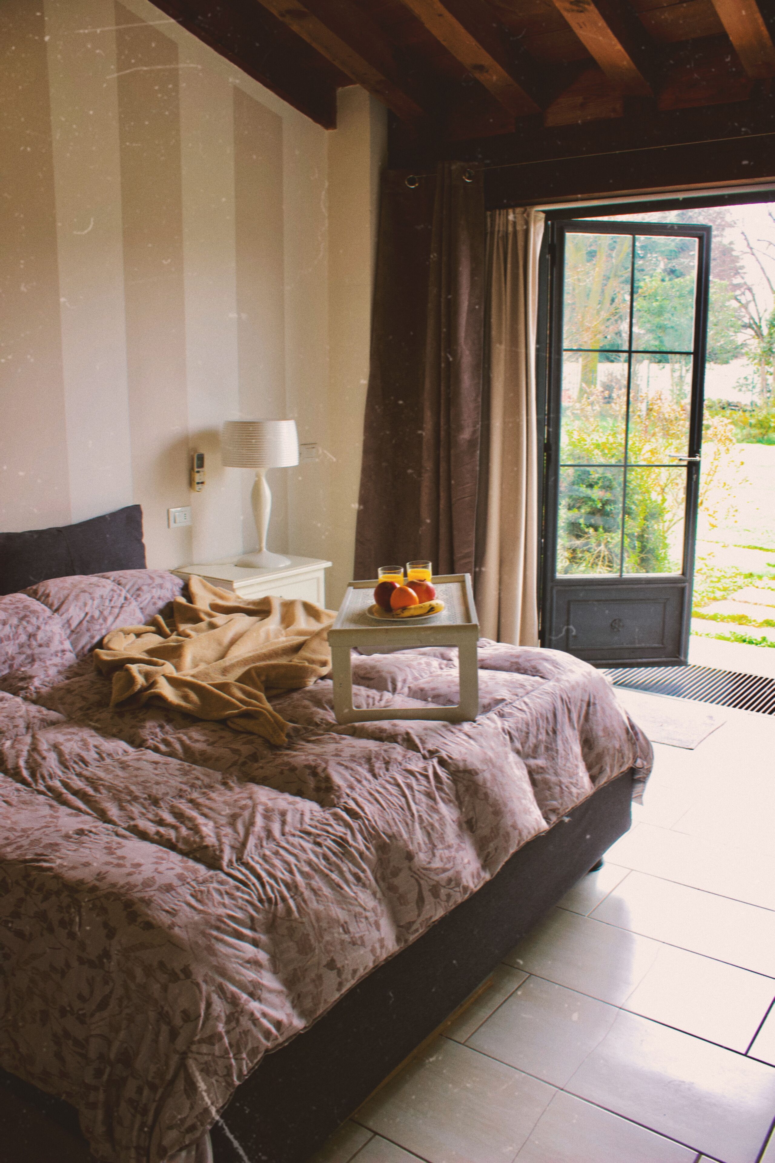 A bedroom with a bed, a bedside table with a lamp, a window with curtains, and a view of greenery outside.
