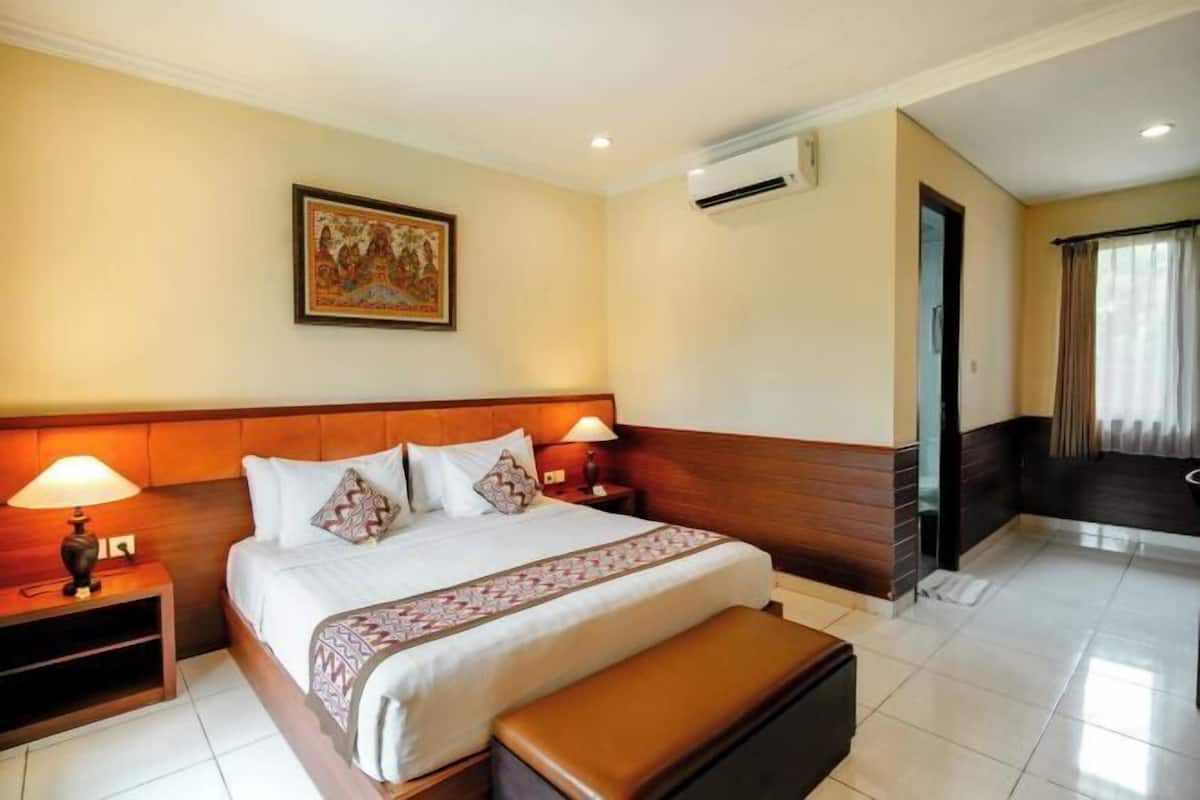 A hotel room with a large bed, two bedside tables with lamps, a wall-mounted air conditioner, a framed picture, and a view of the outside through a window.