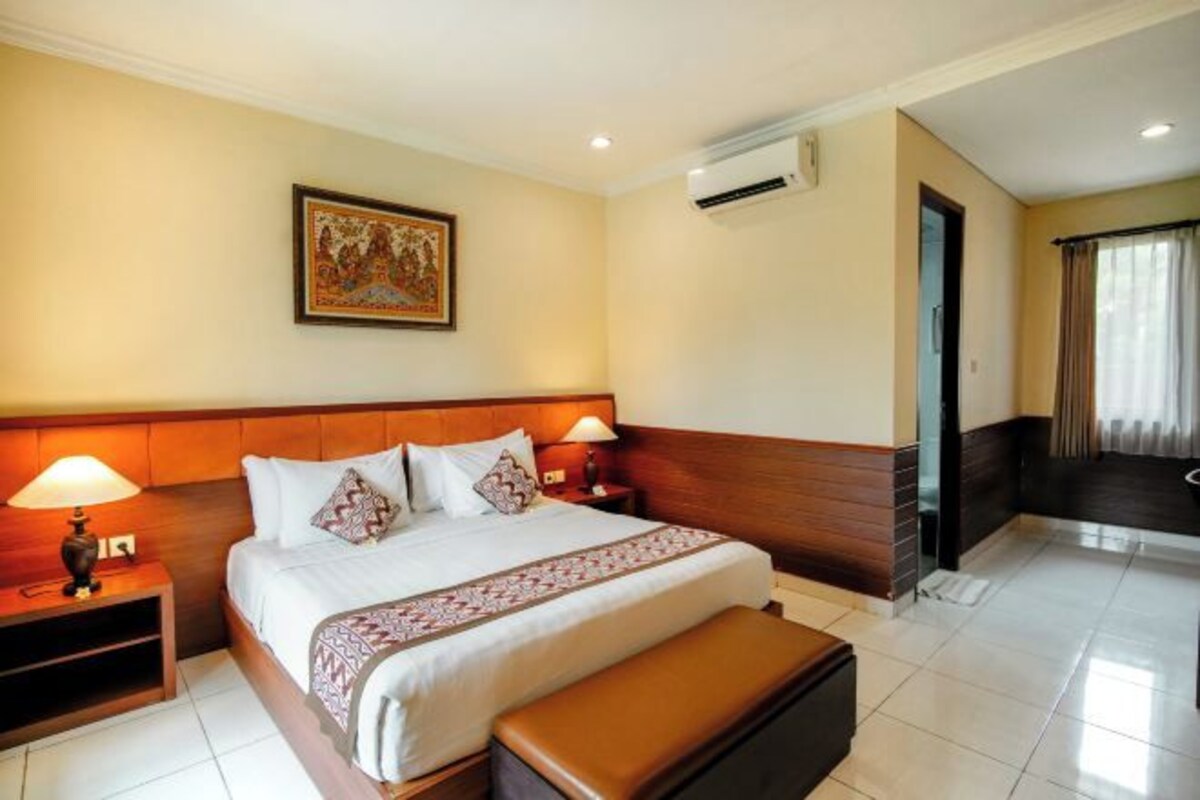 A hotel room with a large bed, two bedside tables with lamps, a wall-mounted air conditioner, a framed picture, and a view of the outside through a window.