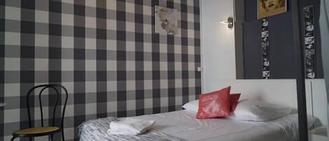 A bedroom with a checkered wall, a bed with white and red pillows, a chair, and a wall-mounted artwork.
