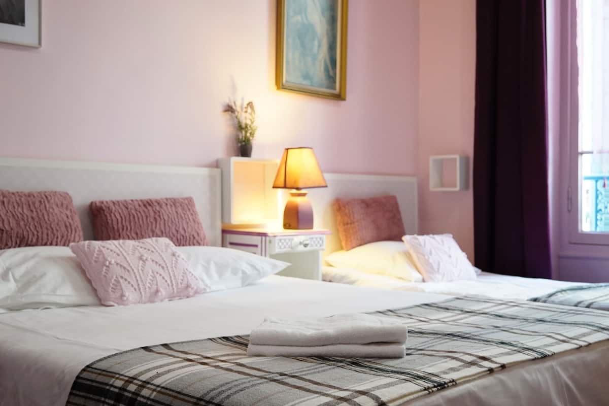 A hotel room with a bed, a nightstand with a lamp, a painting on the wall, and a window with curtains.