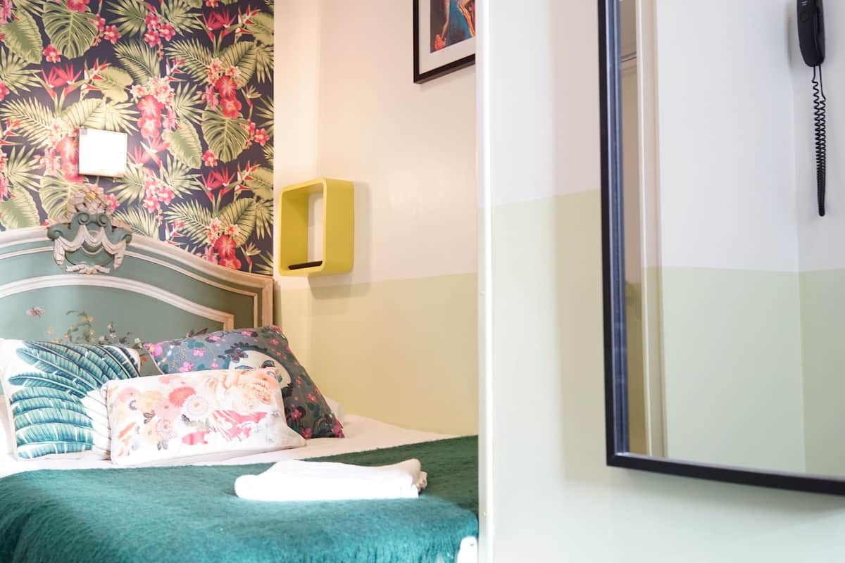 A neatly made bed with floral bedding and a green throw blanket. A small bedside table with a yellow lamp is visible.