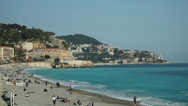 Une plage où des gens se détendent et se promènent le long du rivage, avec des bâtiments en arrière-plan et un ciel dégagé.