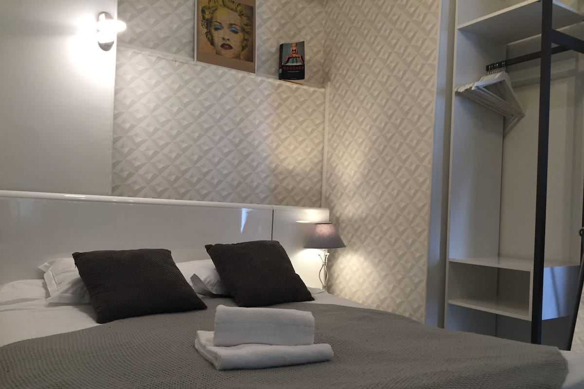 A neatly made bed with a grey blanket and two dark pillows, a bedside lamp, a framed picture on the wall, and a wardrobe with hanging clothes.
