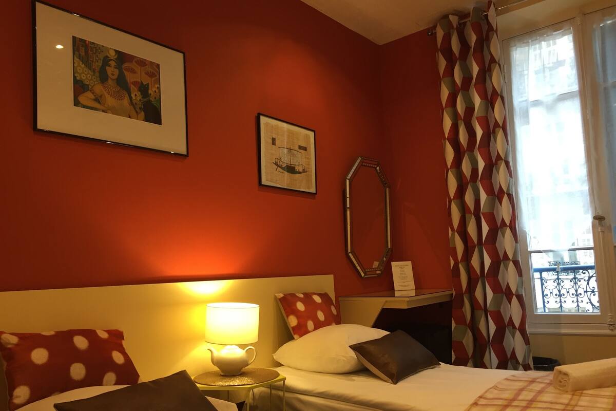 A hotel room with two beds, a nightstand with a lamp, a mirror, and two framed pictures on the wall.