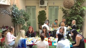 Un groupe de personnes est assis à une table entourée de chaises, en train de dîner en plein air, dans un cadre verdoyant, avec un bâtiment aux volets fermés en arrière-plan.