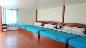 Una habitación con cuatro camas individuales alineadas, cabeceros de madera, paredes blancas y una ventana con cortinas azules.