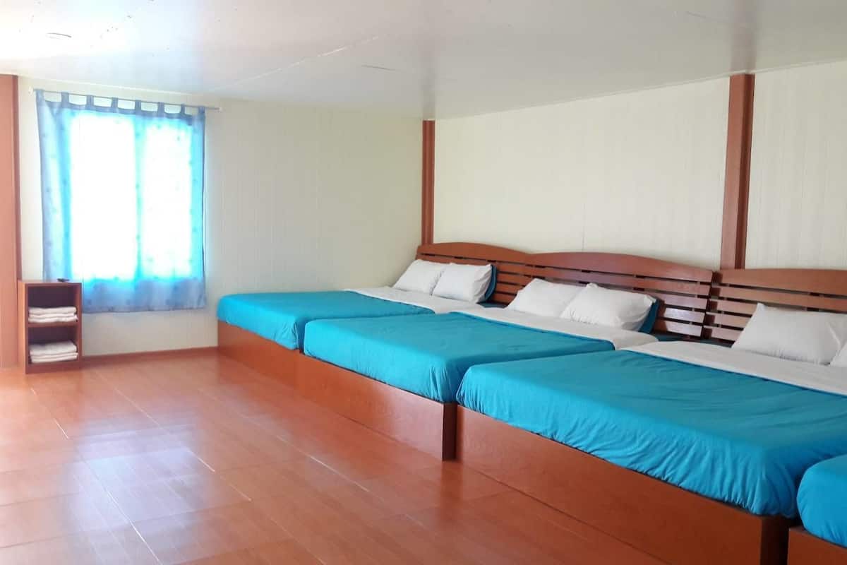 Quarto com quatro camas individuais alinhadas, cabeceiras de madeira, paredes brancas e uma janela com cortinas azuis.