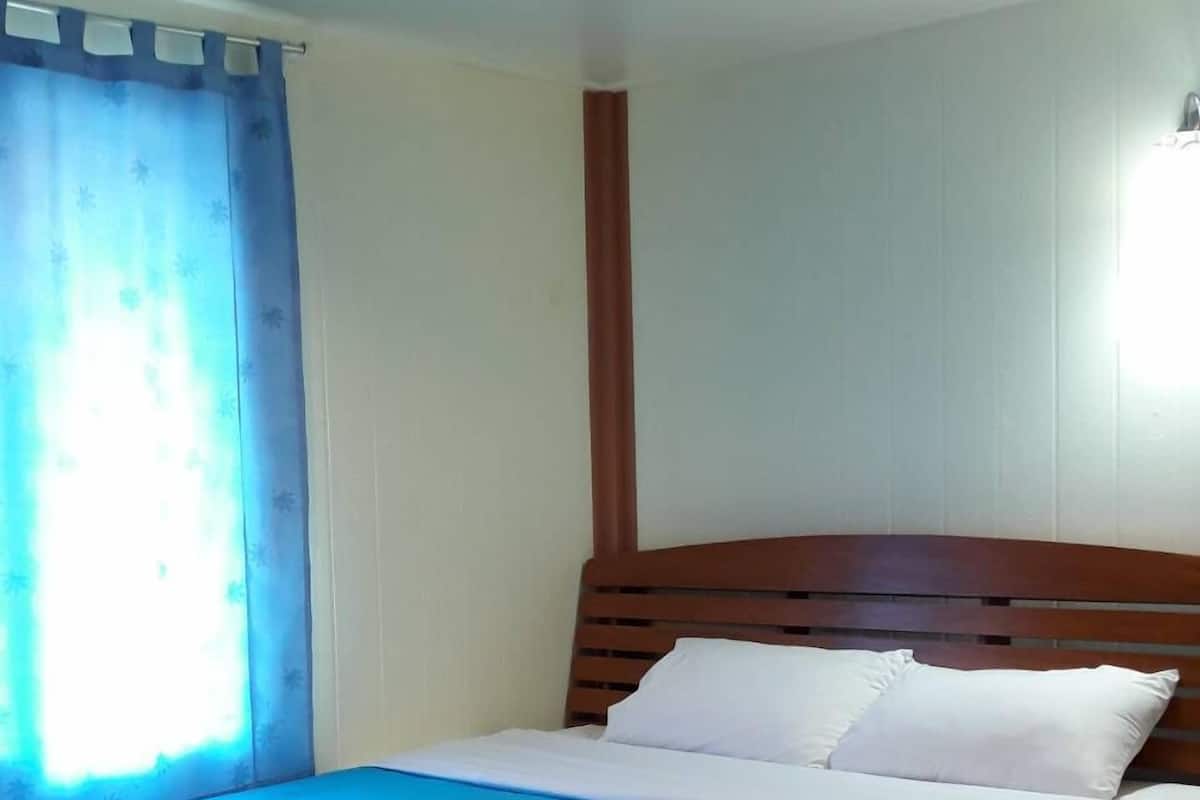 Uma cama de solteiro com cabeceira de madeira, colcha azul e almofadas brancas num quarto com soalho de madeira e janela com cortinas azuis.