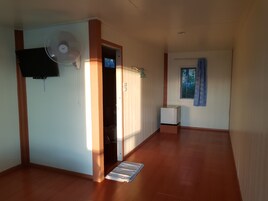 Un pasillo con suelo de madera, un ventilador blanco instalado en la pared, un televisor, una ventana con cortina azul y una puerta que da a otra habitación.