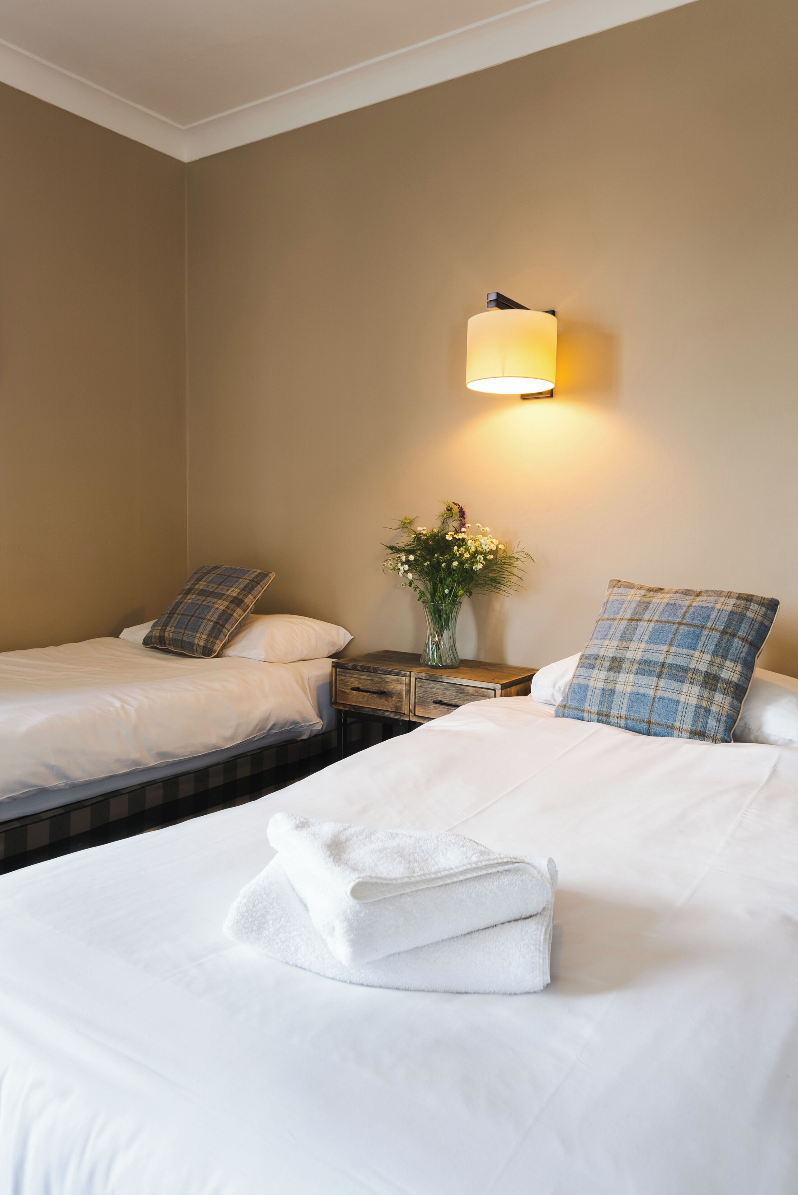A hotel room with two beds, a nightstand, a lamp, and a vase with flowers.