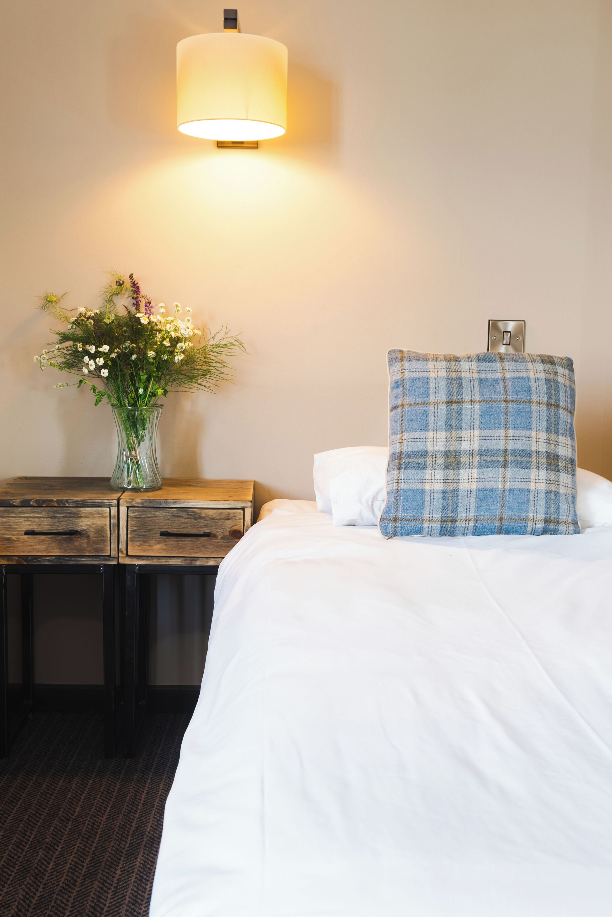 A hotel room with a bed, a nightstand with a vase of flowers, and a wall-mounted lamp.