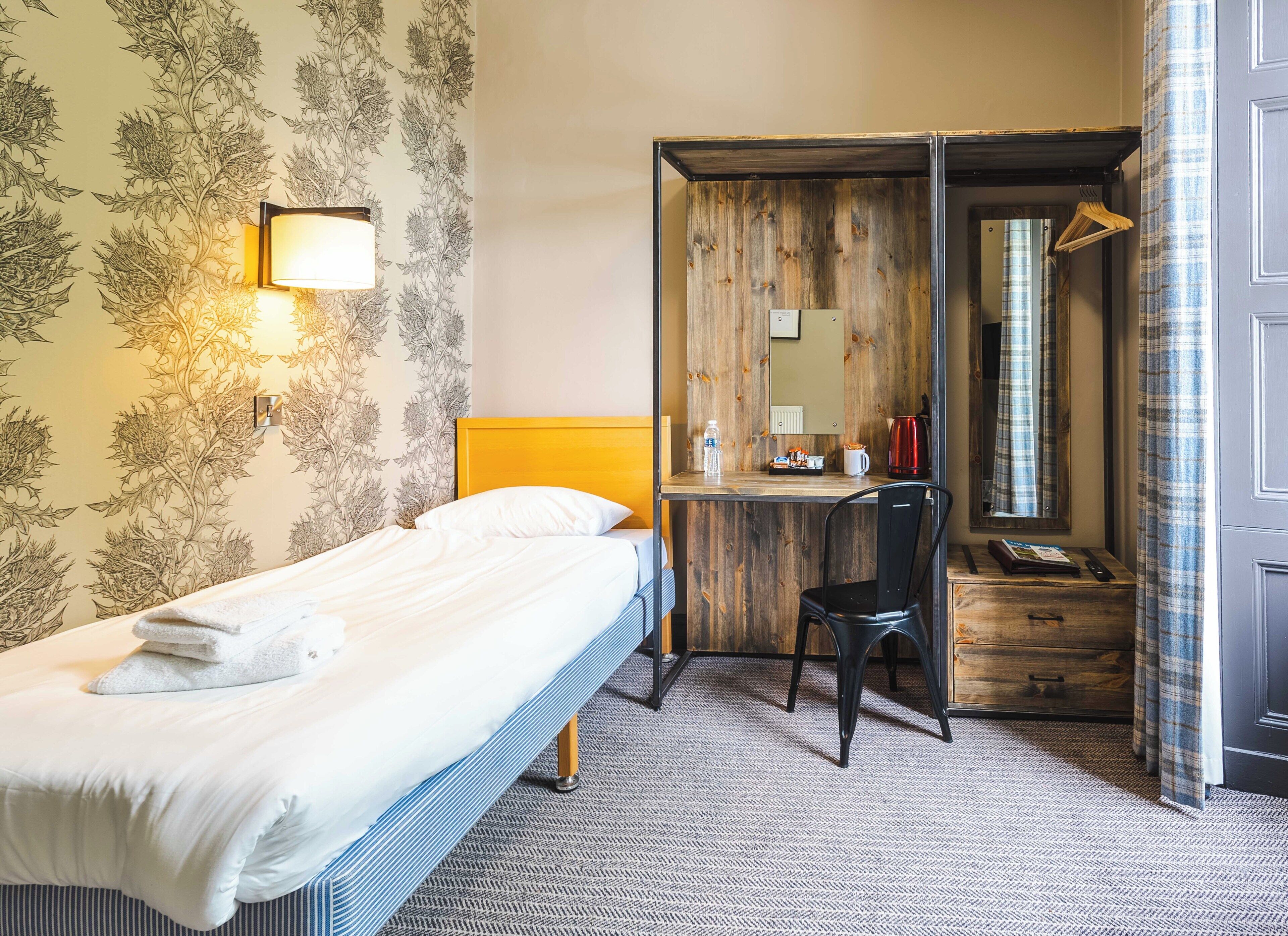 A hotel room with a bed, a desk with a chair, a mirror, and a wardrobe.