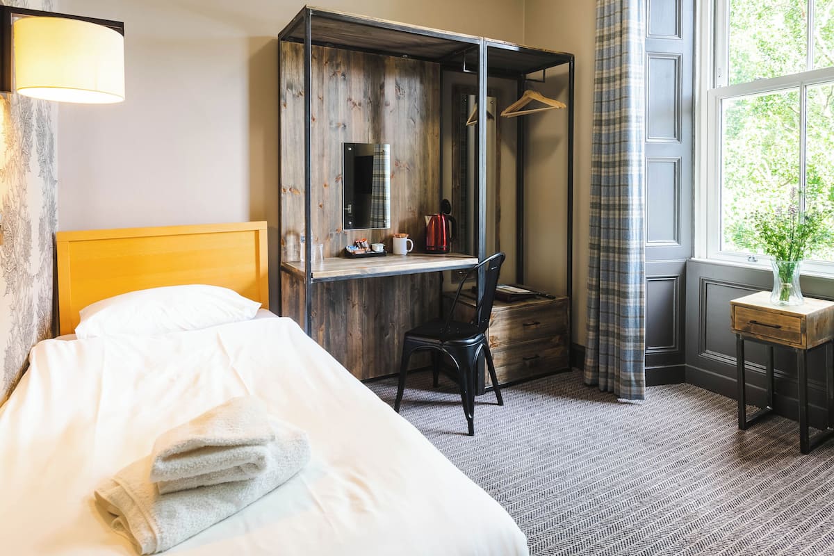 A hotel room with a bed, a desk with a chair, a hanging coat rack, and a window with curtains.
