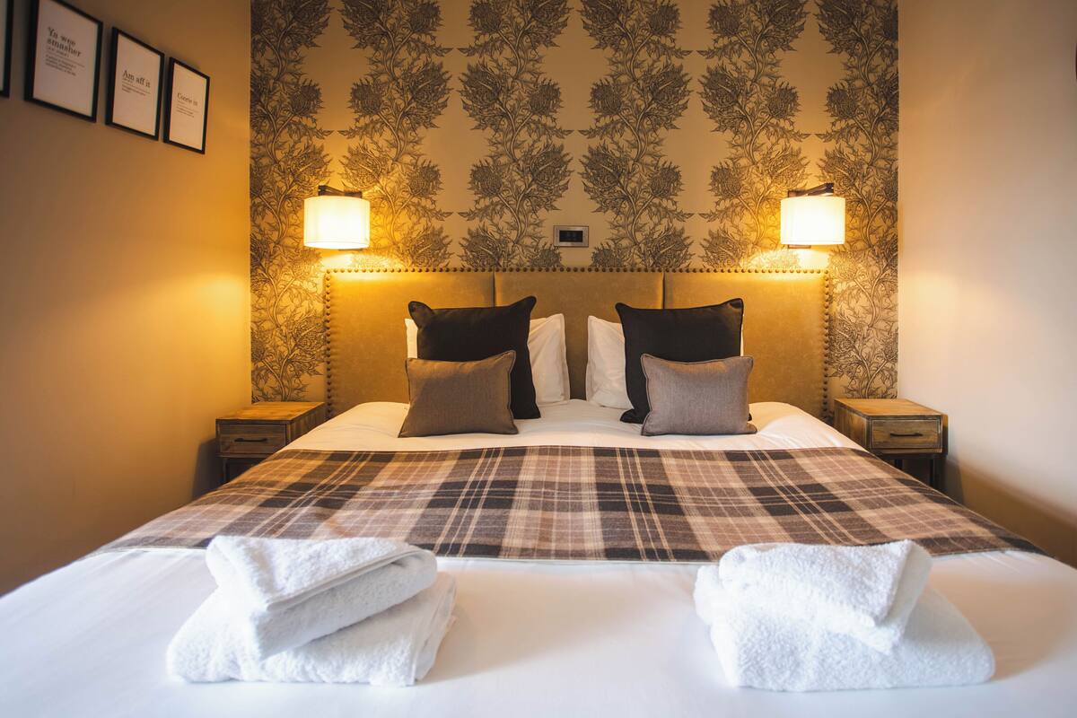 A neatly made bed with a plaid bedspread, two bedside lamps, and a floral wallpaper pattern.