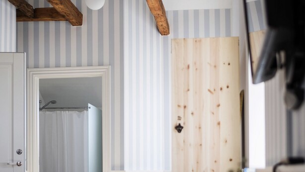 Ein Zimmer mit gestreifter Tapete, Holzbalken, einer weißen Tür und einer Holzbank mit einer Vase.
