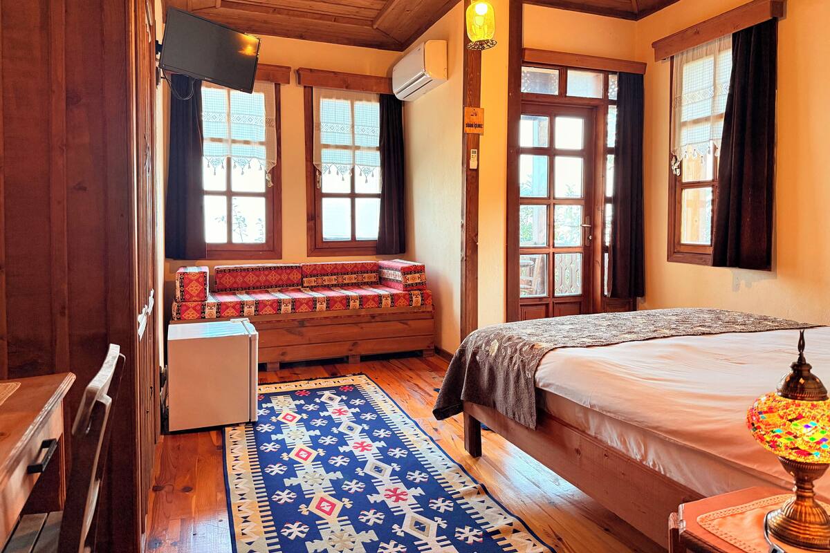 A cozy room with a wooden ceiling, a bed with a patterned bedspread, a bench with cushions, a small table, and a lamp.