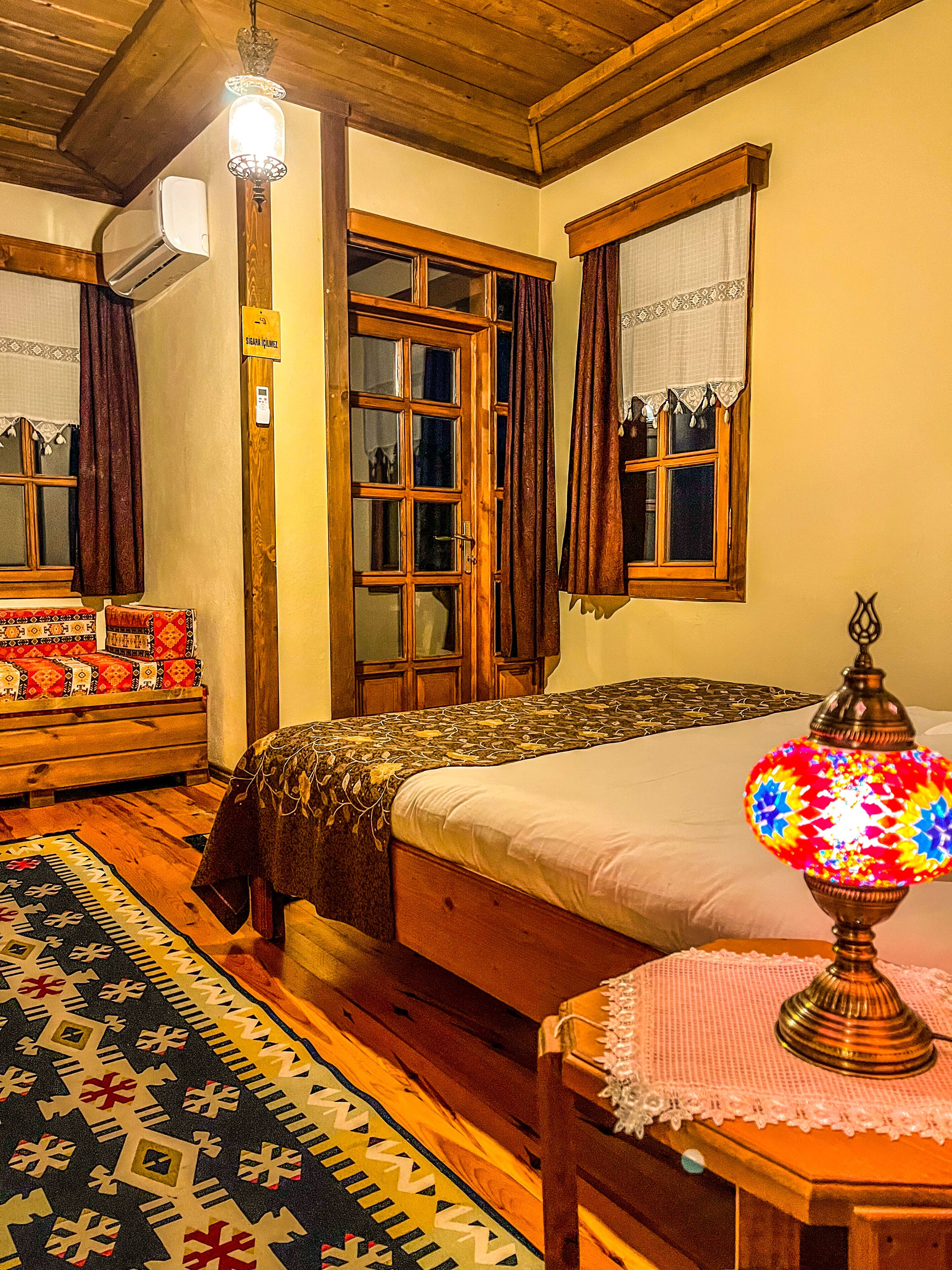 Une chambre traditionnelle avec un lit en bois, un tapis à motifs, une table avec une lampe et une fenêtre avec des rideaux.