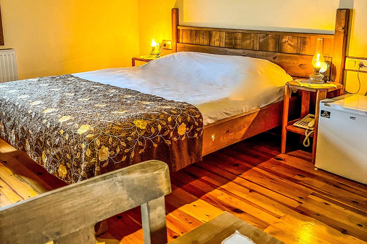 A hotel room with a wooden floor, a bed with a floral bedspread, a nightstand with a lamp, a painting on the wall, and a chair.