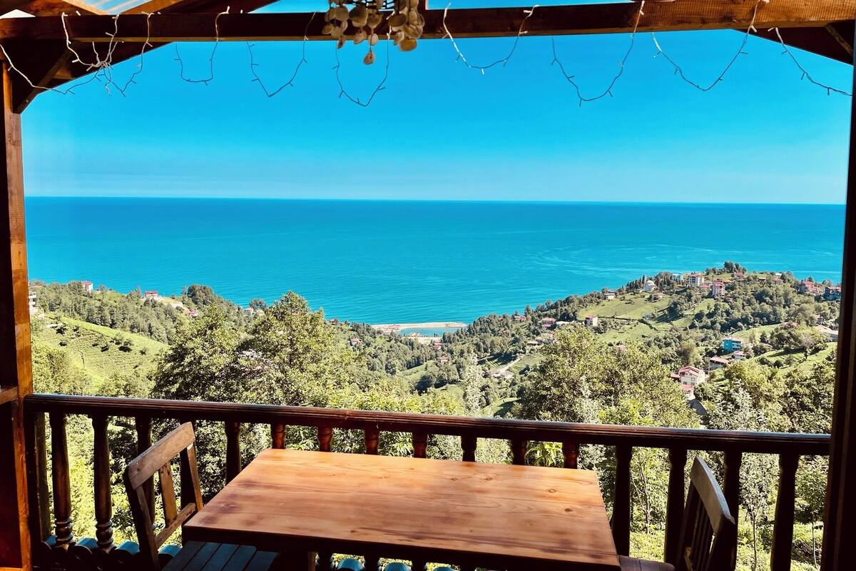 A wooden deck with a table and chairs, overlooking a coastal landscape with a clear view of the sea and hills.