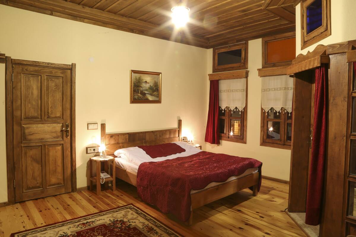 A wooden-floored bedroom with a large bed, a nightstand, a wall-mounted lamp, and a framed picture.