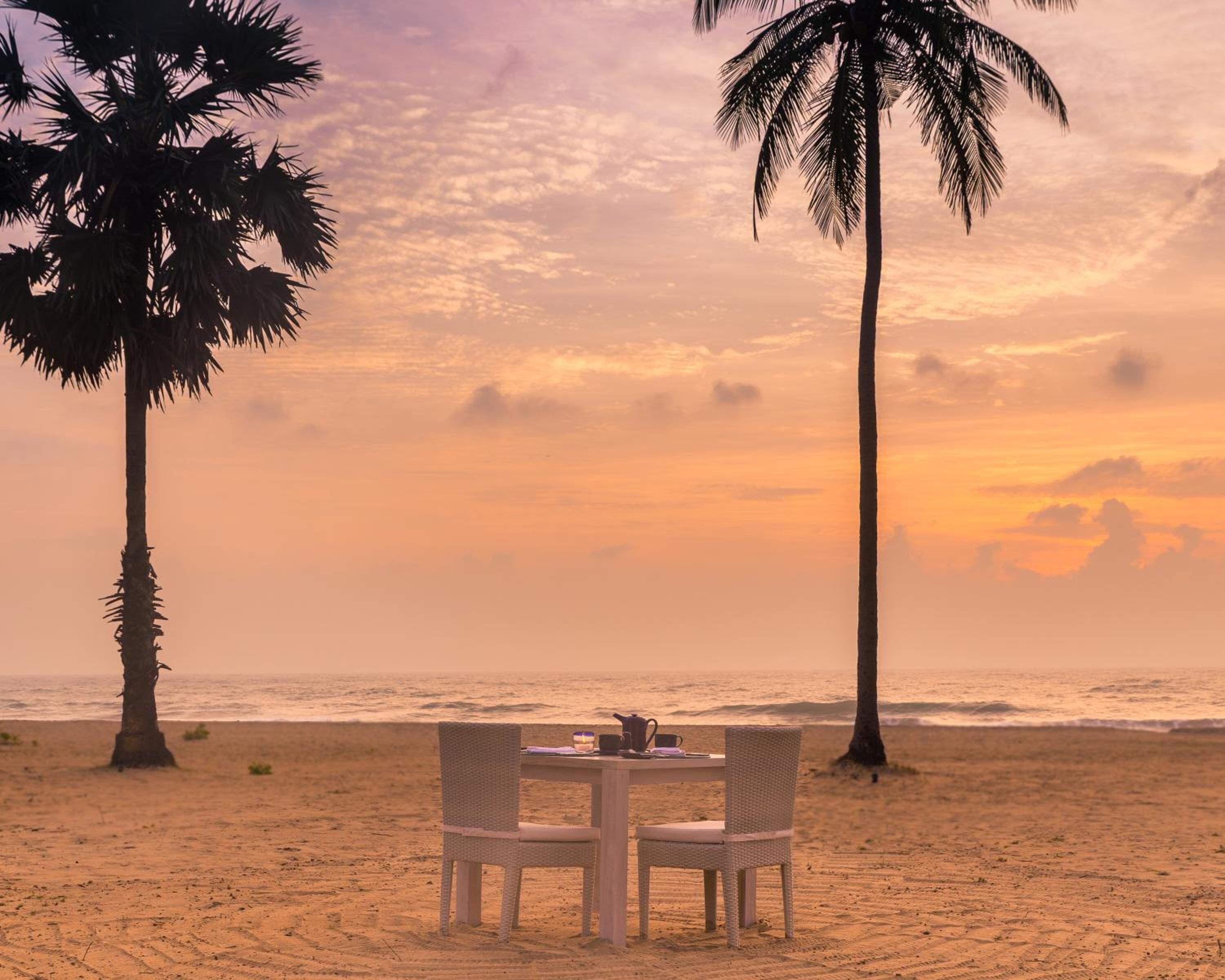 A beach setting with a table and chairs, palm trees, and a sunset view.