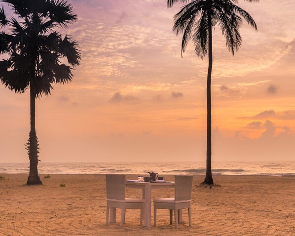 A beach setting with a table and chairs, palm trees, and a sunset view. - Jetwing Surf & Safari, A Luxury Reserve (Pottuvil)