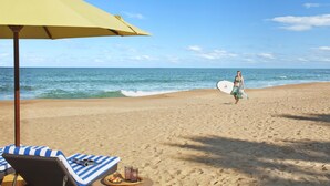 A beach with a yellow umbrella, lounge chairs, and a person carrying a surfboard. - Jetwing Surf & Safari, A Luxury Reserve (Pottuvil)