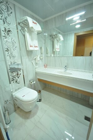 A bathroom with a toilet, sink, and mirror. There are decorative tiles on the walls and a towel rack with towels. - Bupa Hotel (Kayseri)