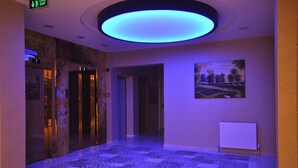 A hallway with a patterned floor, an elevator, a painting on the wall, and a ceiling light. - Bupa Hotel (Kayseri)
