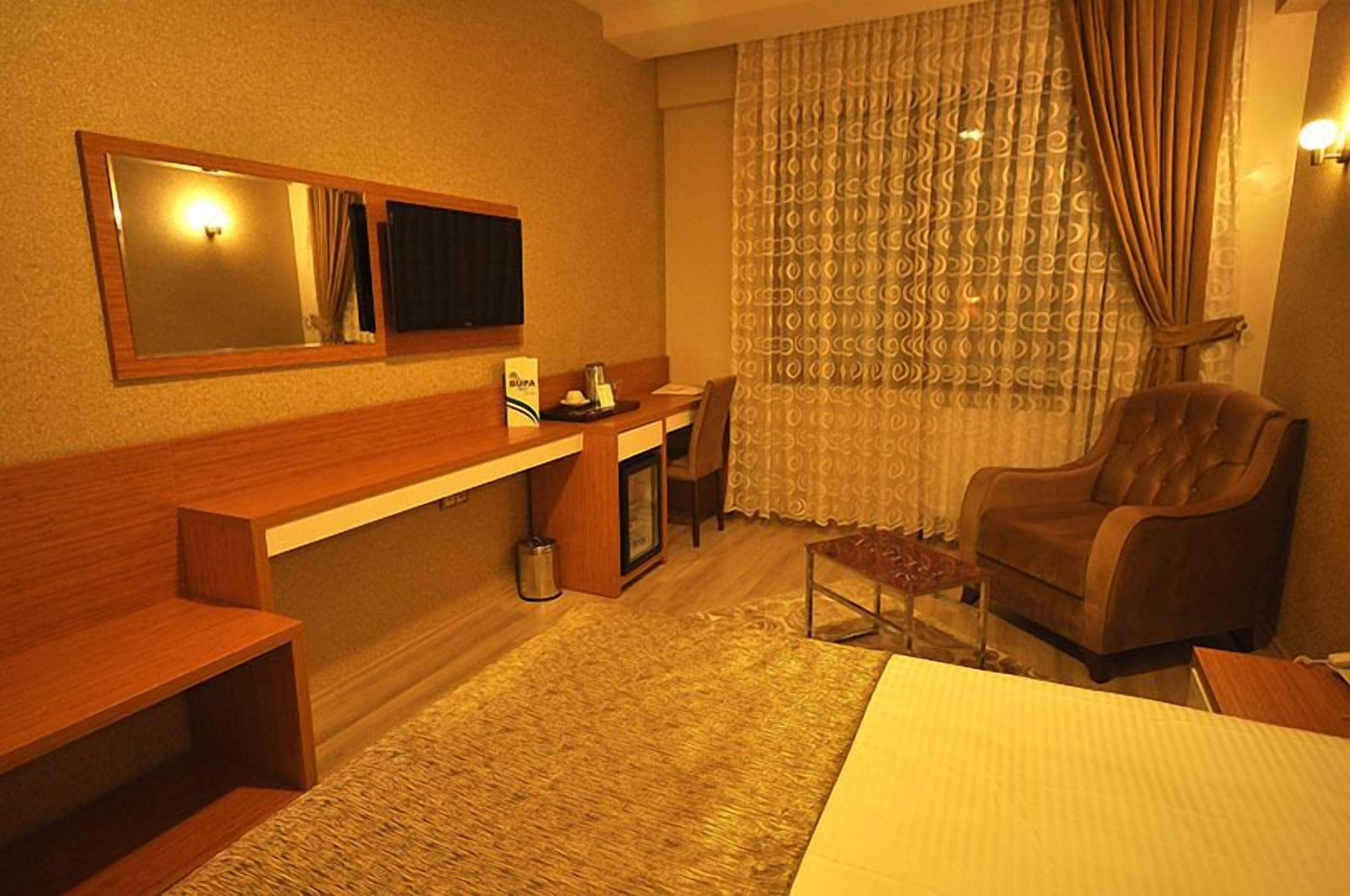 Une chambre d’hôtel avec un banc en bois, une petite table, une chaise, un lit, un bureau, un miroir et une fenêtre avec des rideaux à motifs.