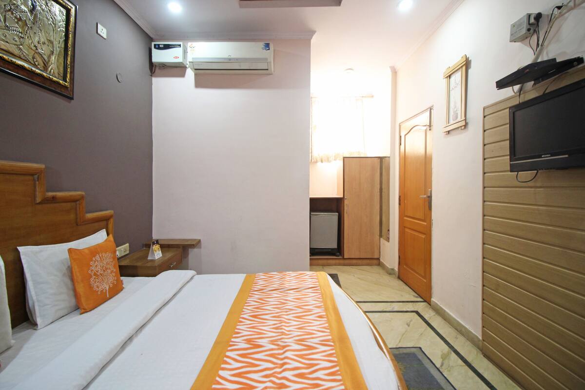 A hotel room with a bed, a nightstand, a TV mounted on the wall, and an air conditioning unit.