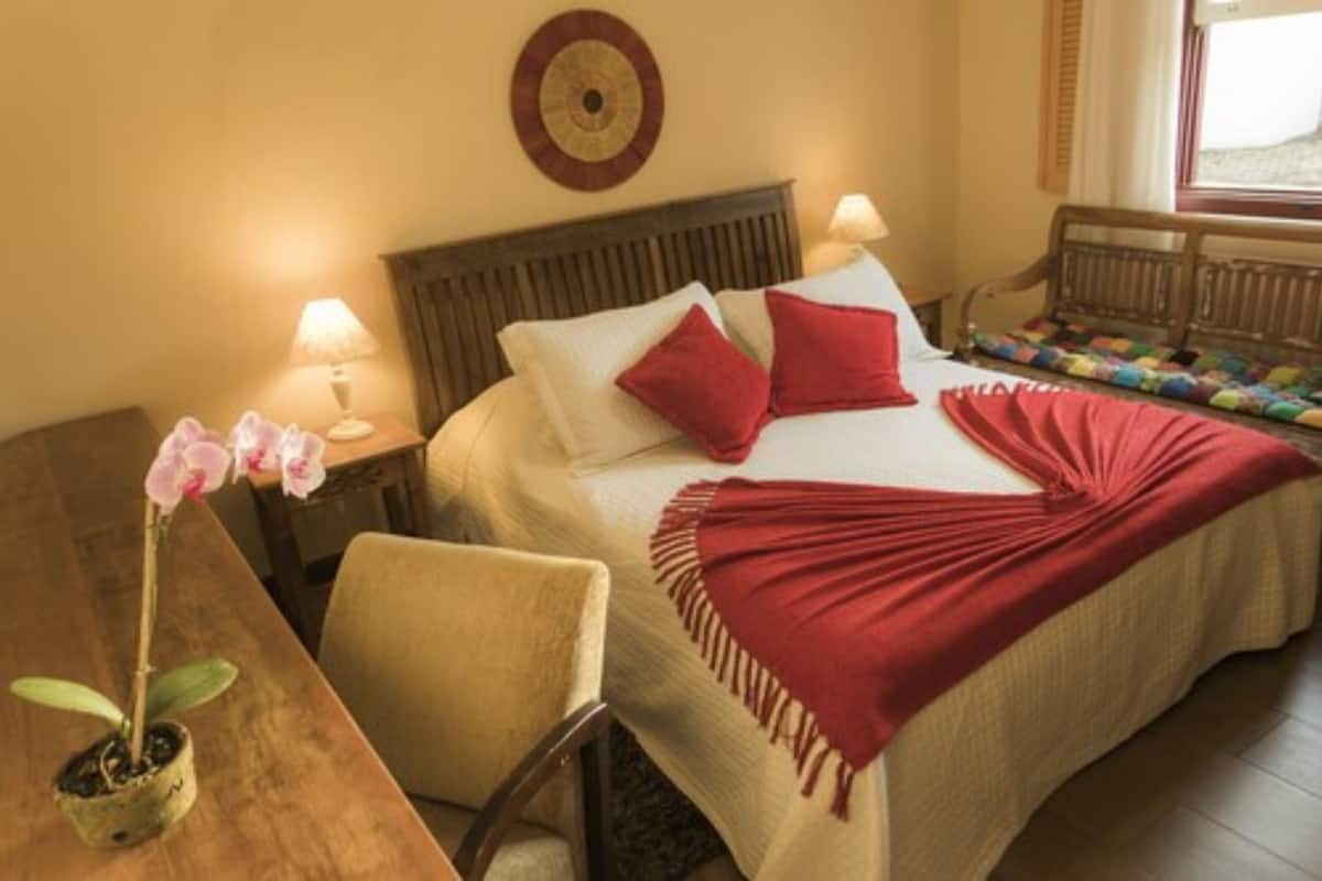 A hotel room with a bed, a wooden nightstand, a chair, a desk, a potted orchid, and a window with curtains.