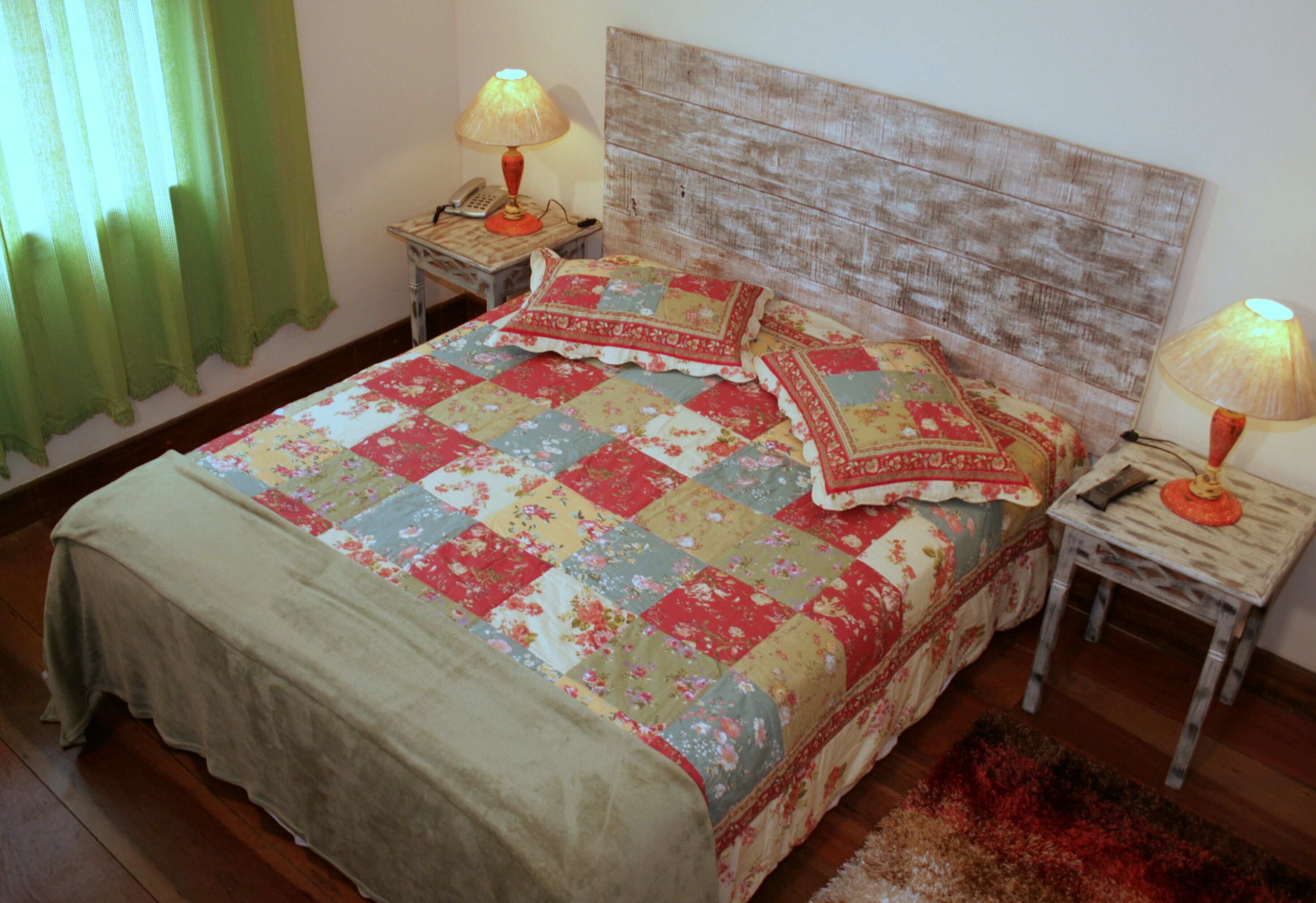 A bed with a floral quilt, two bedside lamps, a wooden headboard, and a green curtain.