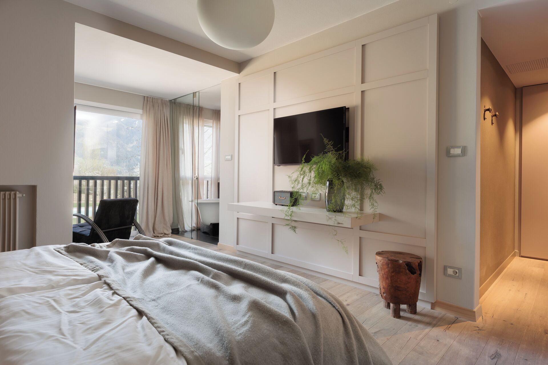 Ein modernes Schlafzimmer mit einem großen Bett, einem an der Wand befestigten Fernseher, einer kleinen Pflanze und einem Holzschemel.