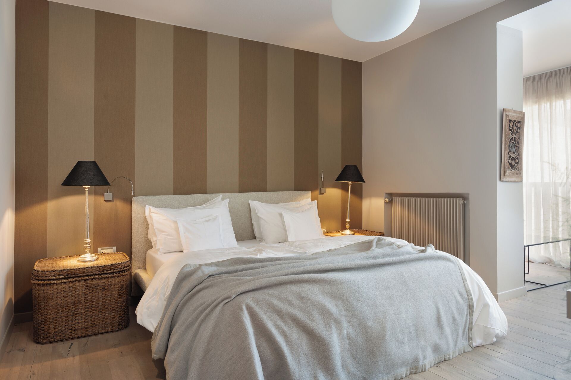 A neatly made bed with white linens, two bedside lamps, a wicker side table, and a radiator.