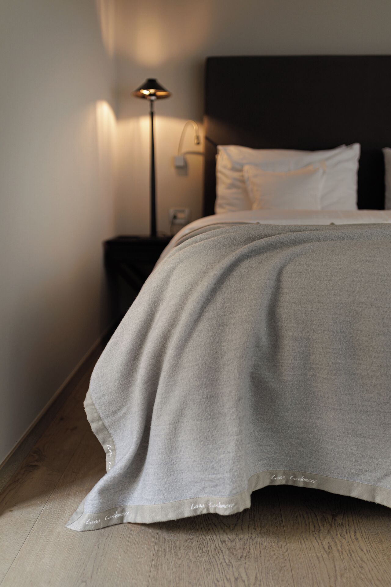 A neatly made bed with a grey blanket, a bedside lamp, and a dark headboard.