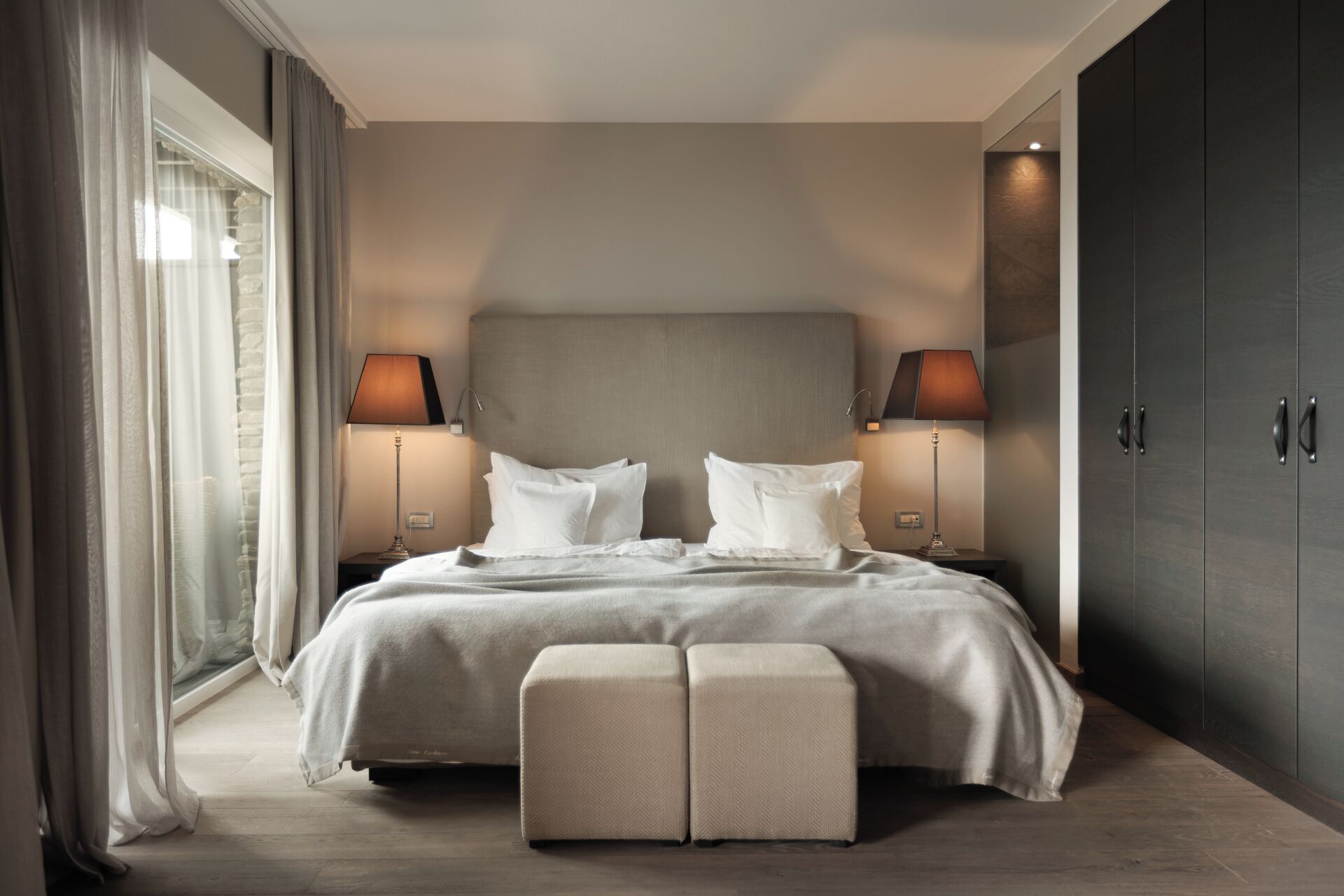 A modern bedroom with a large bed, two bedside tables with lamps, a dark wardrobe, and a large window with curtains.