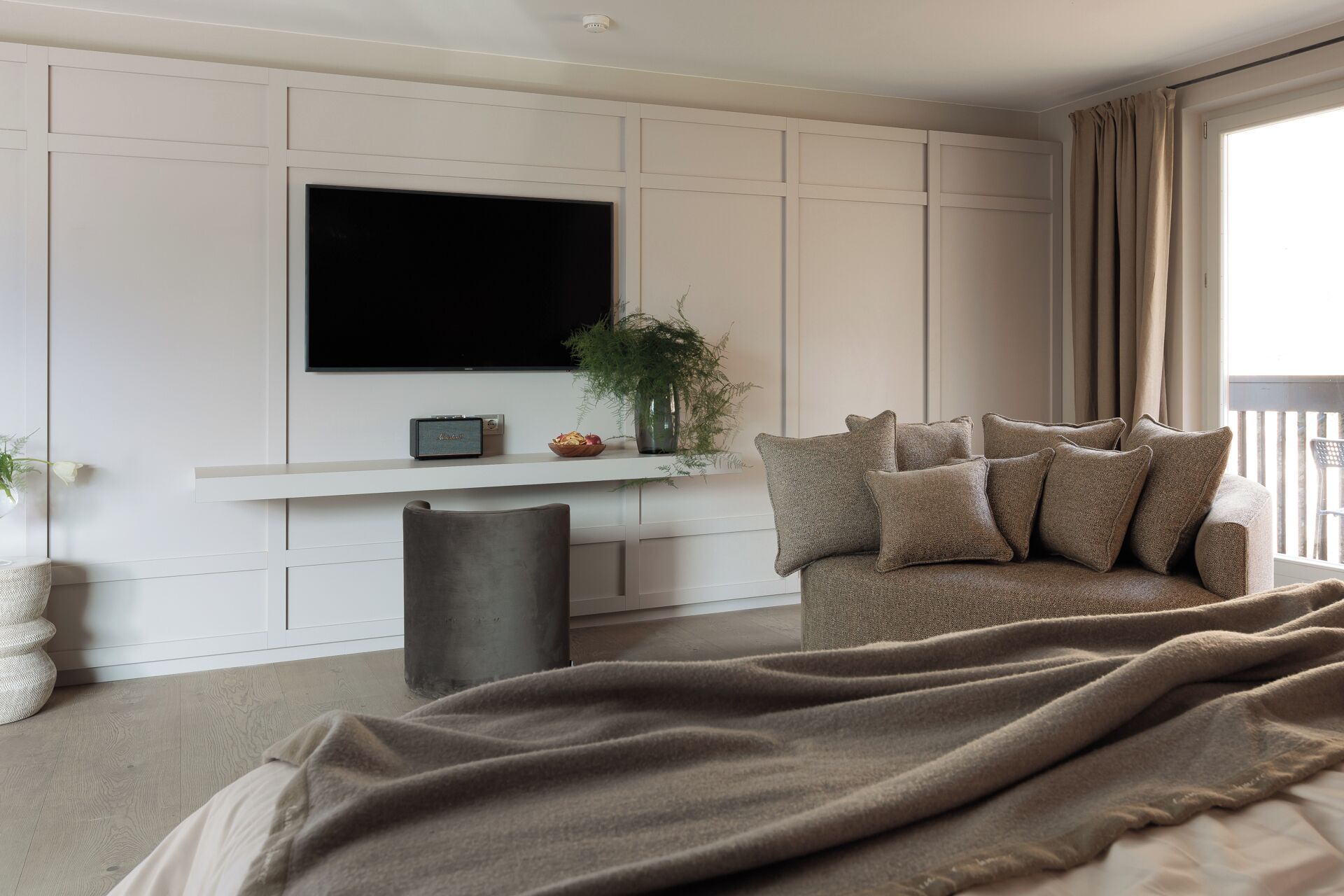 A modern bedroom with a large bed, a sofa, a TV mounted on the wall, and a small plant on a shelf.