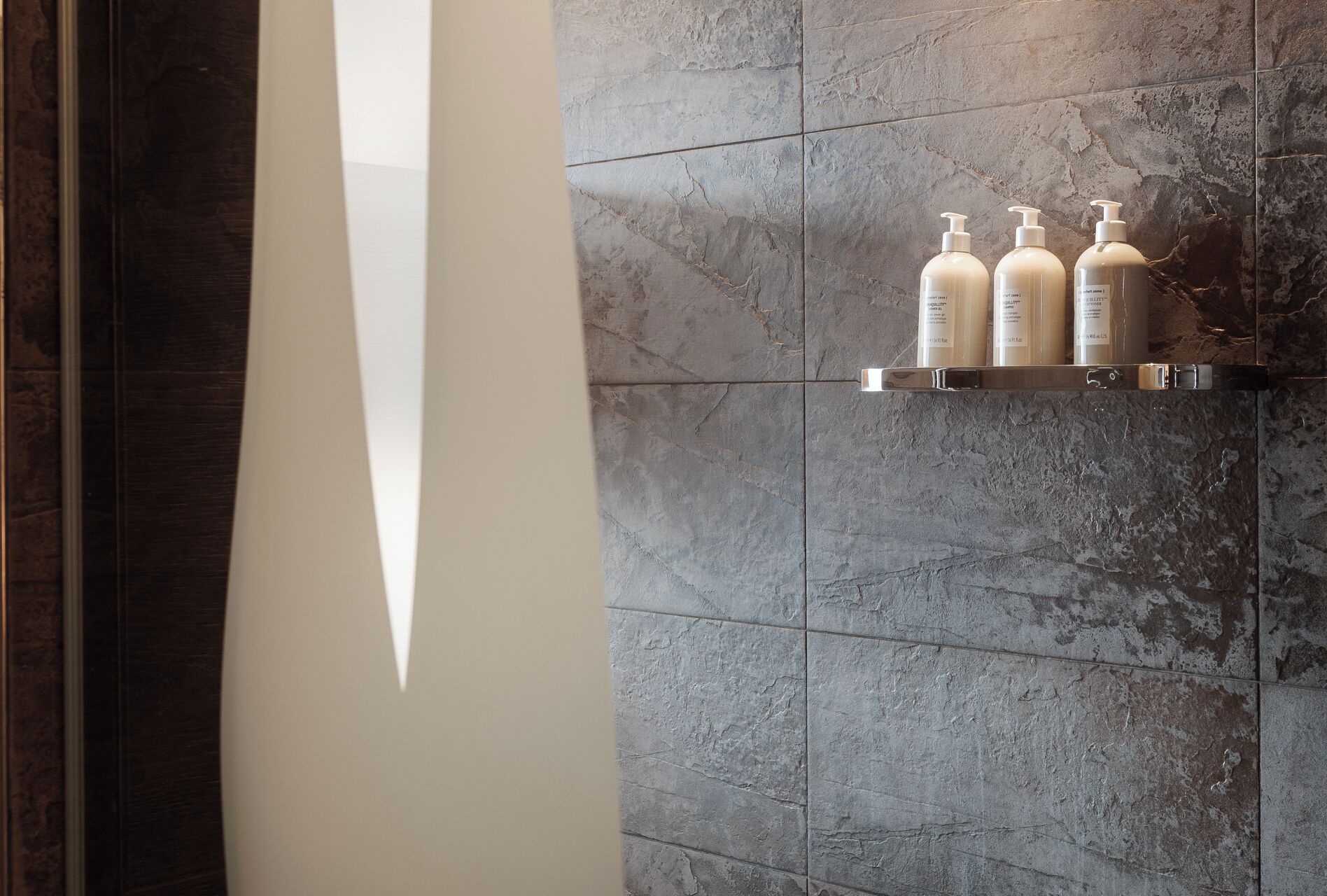 Un baño con baldosas de piedra gris, una pared blanca y un estante con tres botellas de artículos de aseo.