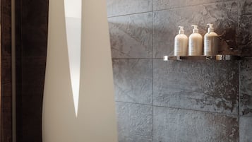 Un baño con baldosas de piedra gris, una pared blanca y un estante con tres botellas de artículos de aseo.