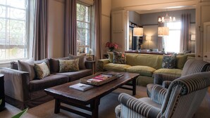 A cozy living room with a brown leather sofa, a green sofa, and a striped armchair. There is a wooden coffee table with books and a vase of flowers. Large windows with curtains allow natural light to fill the room.